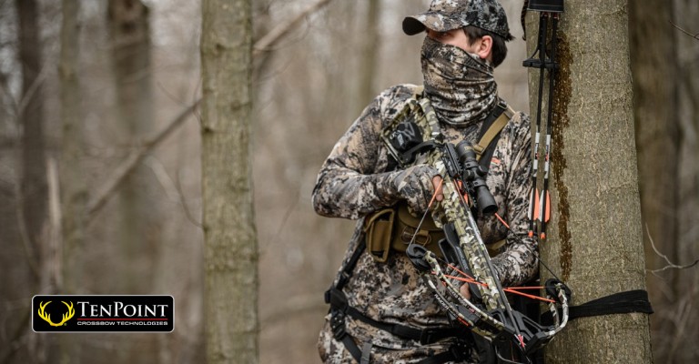Hunter holding a crossbow next to a tree in the woods
