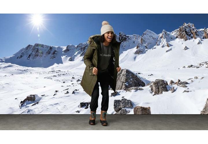 Woman standing in front of a sunny, snowy moutain backdrop wearing the Columbia Suttle Mountain Jacket smiling